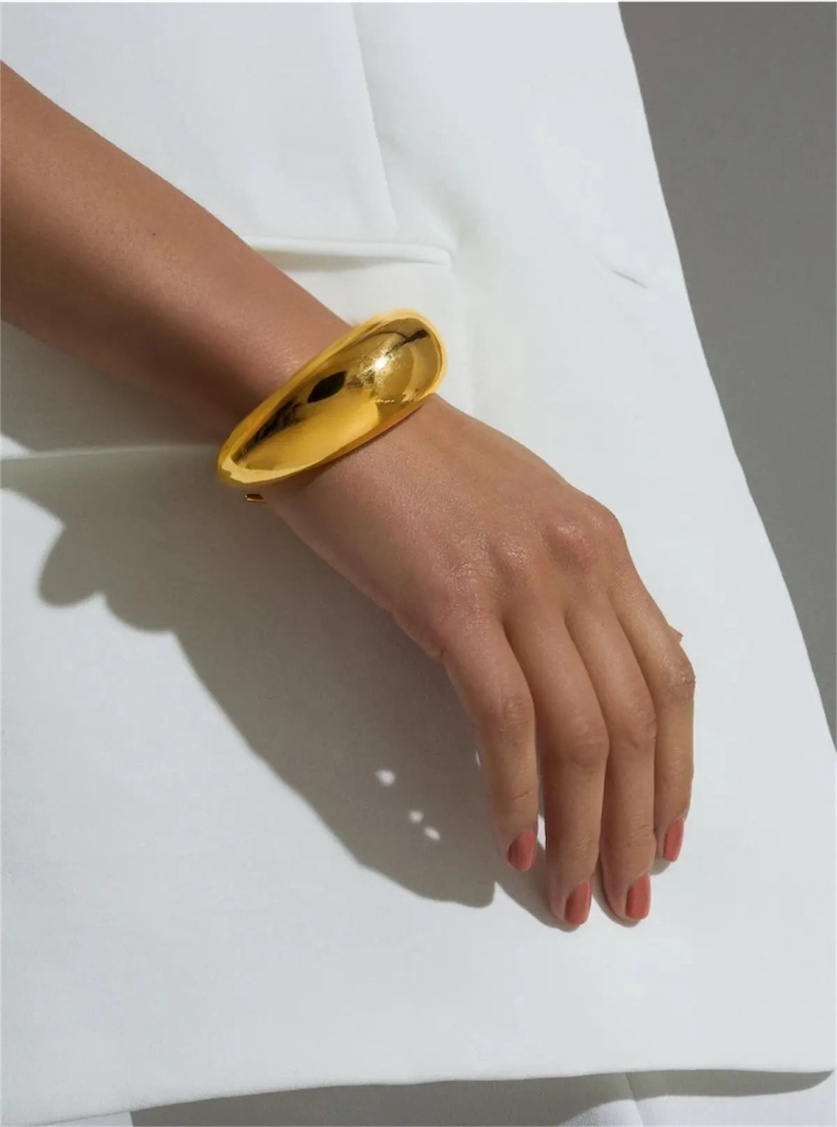 A person with light skin tone wears the wide, shiny AEQEA LUNA CUFF gold bracelet on their wrist. Their hand rests on a white garment against a neutral background, and their nails are neatly manicured with a natural finish.