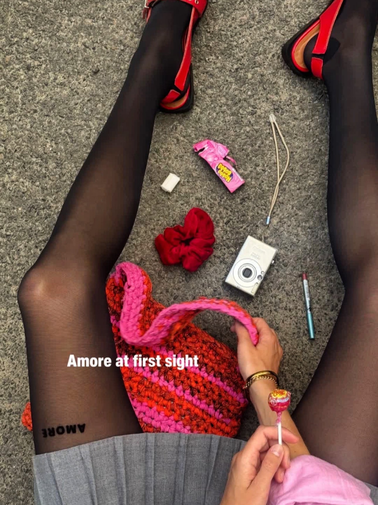 Wearing AEQEA AMORE TIGHTS in black (40 DEN), a person sits on the ground in red sandals and a gray skirt. Nearby are a red scrunchie, chewing gum, camera, lipstick, pink knit bag, and lollipop. Text: "Amore at first sight.