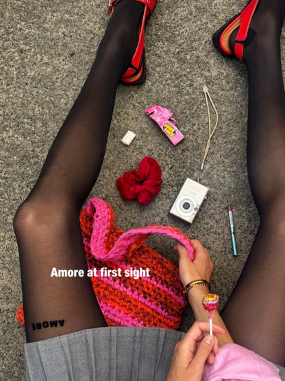 Wearing AEQEA AMORE TIGHTS in black (40 DEN), a person sits on the ground in red sandals and a gray skirt. Nearby are a red scrunchie, chewing gum, camera, lipstick, pink knit bag, and lollipop. Text: "Amore at first sight.