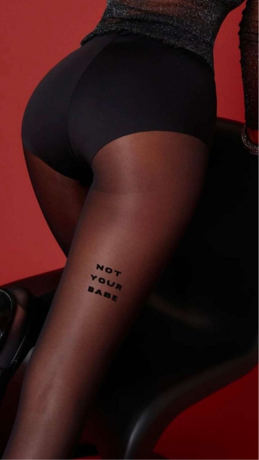 Wearing AEQEA's NOT YOUR BABE TIGHTS—sheer black with "NOT YOUR BABE" on the thigh—a person sits on a black chair, styled with black shorts against a red backdrop.