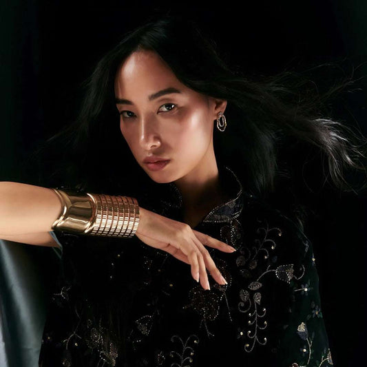 Model wearing CAGE CUFF in gold bronze, showcasing flexible geometric design against dark velvet clothing