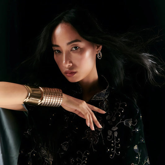 Model wearing CAGE CUFF in gold bronze, showcasing flexible geometric design against dark velvet clothing