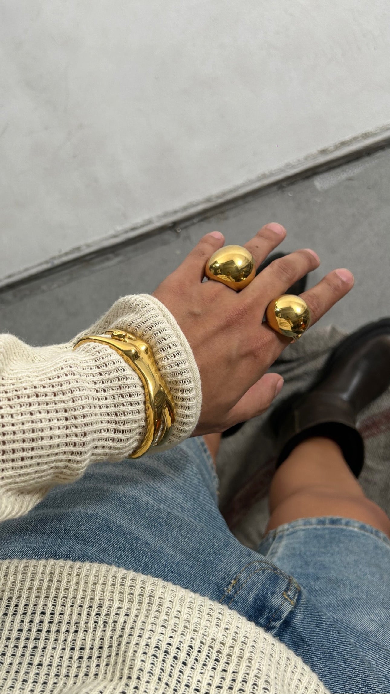 Seen from above, a person in a cream knit sweater and denim shorts wears brown shoes on a patterned floor. Their hand is adorned with chunky 22k gold plated rings and the handcrafted AEQEA EMBRACE CUFF bracelet.