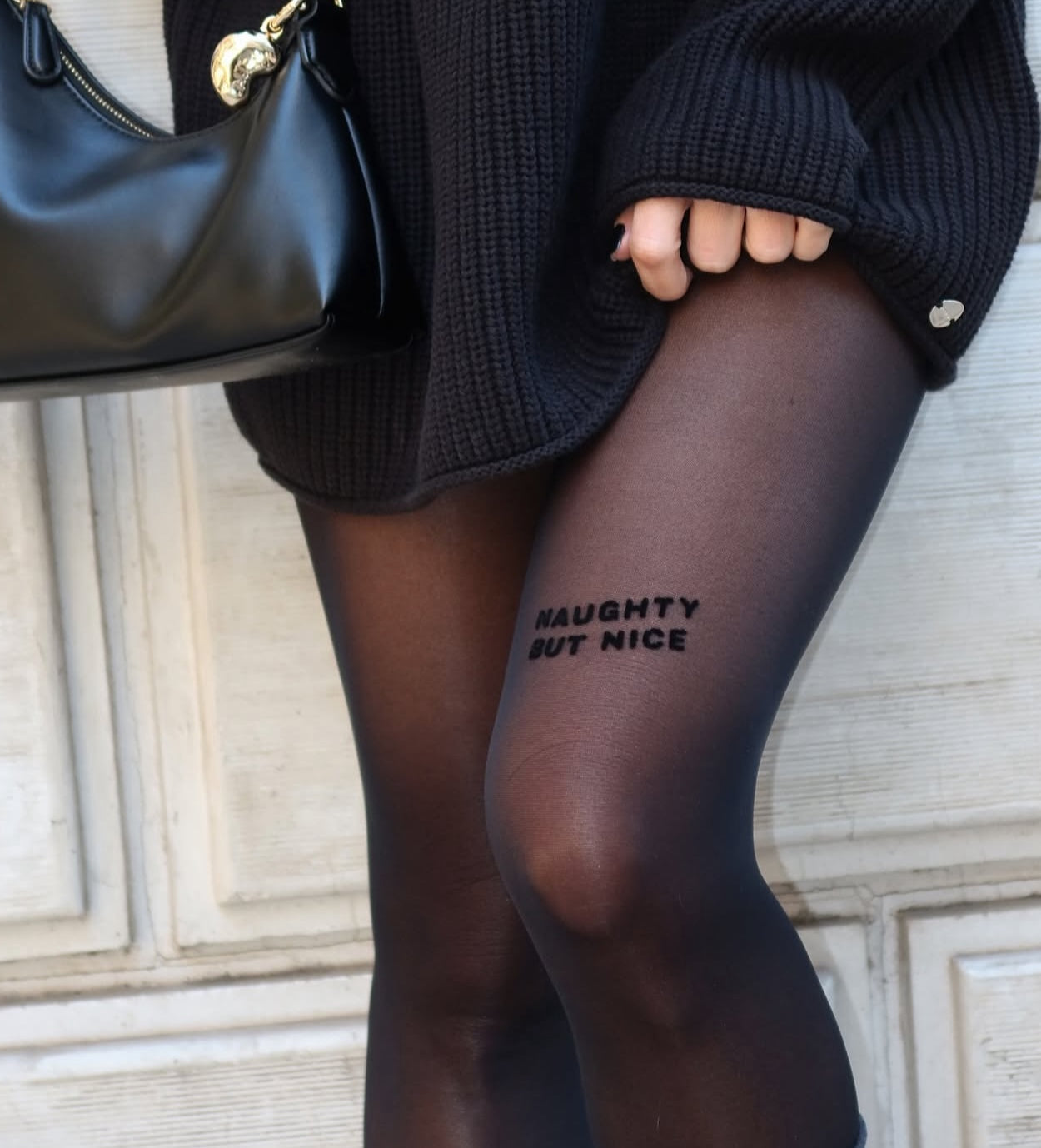 A person in AEQEA’s NAUGHTY BUT NICE TIGHTS and a dark sweater stands before a white textured wall, holding a black handbag.