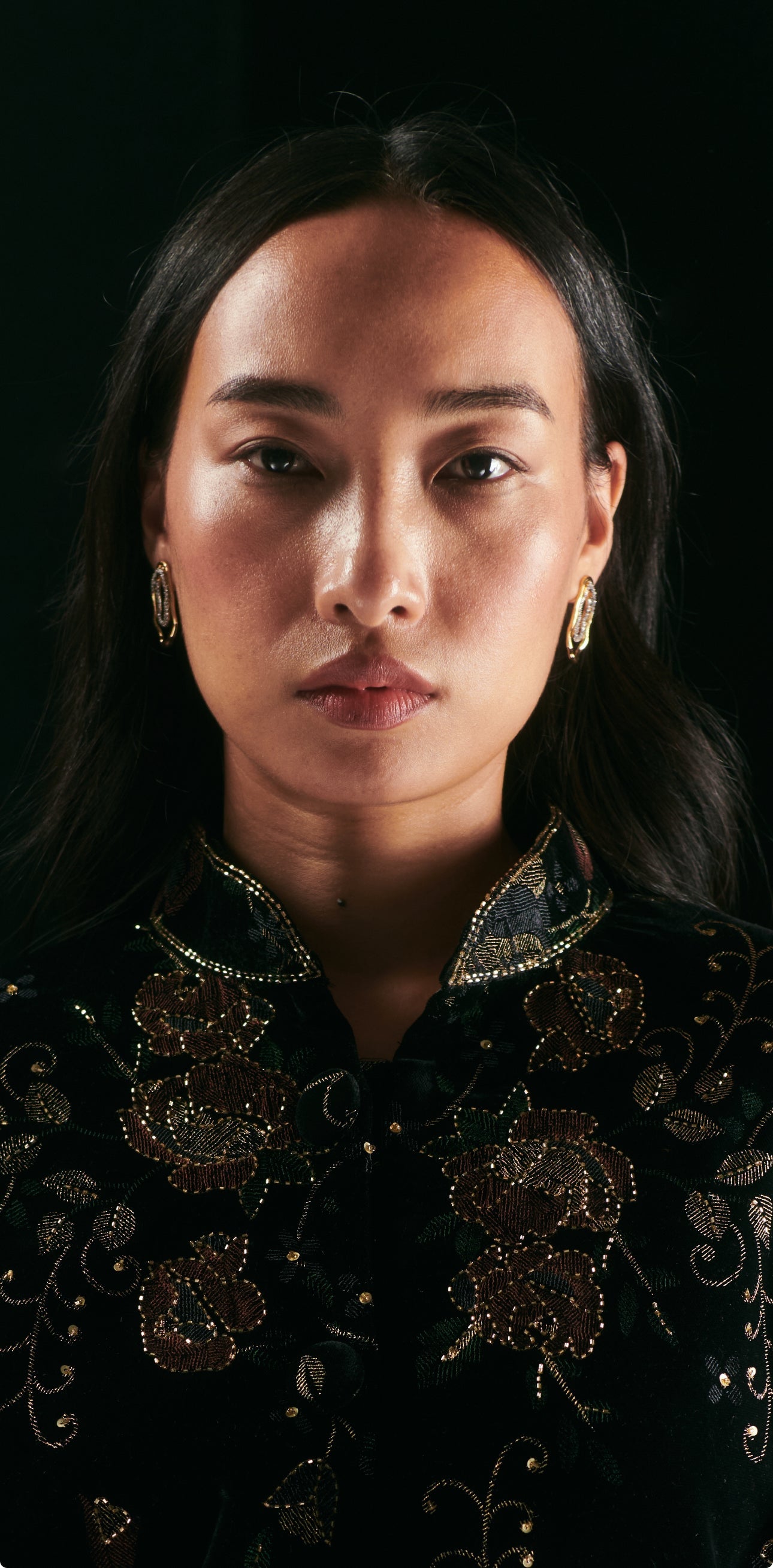 A woman with long dark hair, wearing AEQEA’s SOLANALES CRYSTAL DOUBLE LOOP EARRINGS and a black, gold-embroidered top, faces the camera with a neutral expression against a dark background.