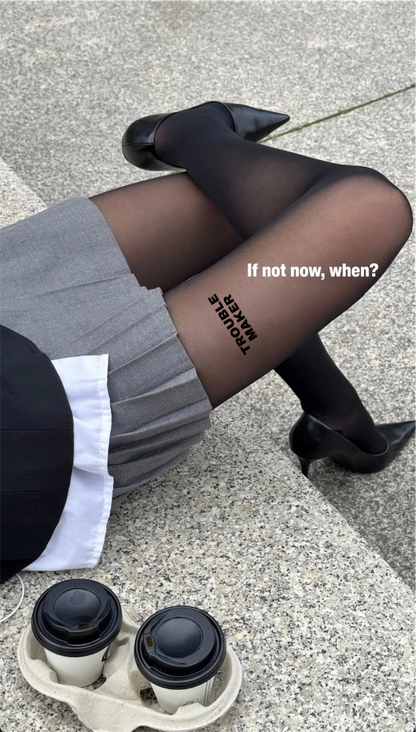 A person in AEQEA TROUBLE MAKER TIGHTS—black statement tights reading "TROUBLE MAKER" and "If not now, when?"—paired with a grey skirt and black shoes, sits on stone beside two takeaway coffee cups.