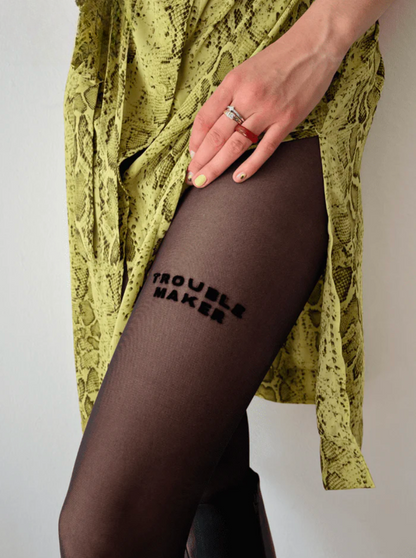A person wears AEQEA's TROUBLE MAKER TIGHTS—black statement tights with "TROUBLE MAKER" on the thigh—paired with a yellow patterned side-slit dress, light polish, and rings for a bold, playful look.