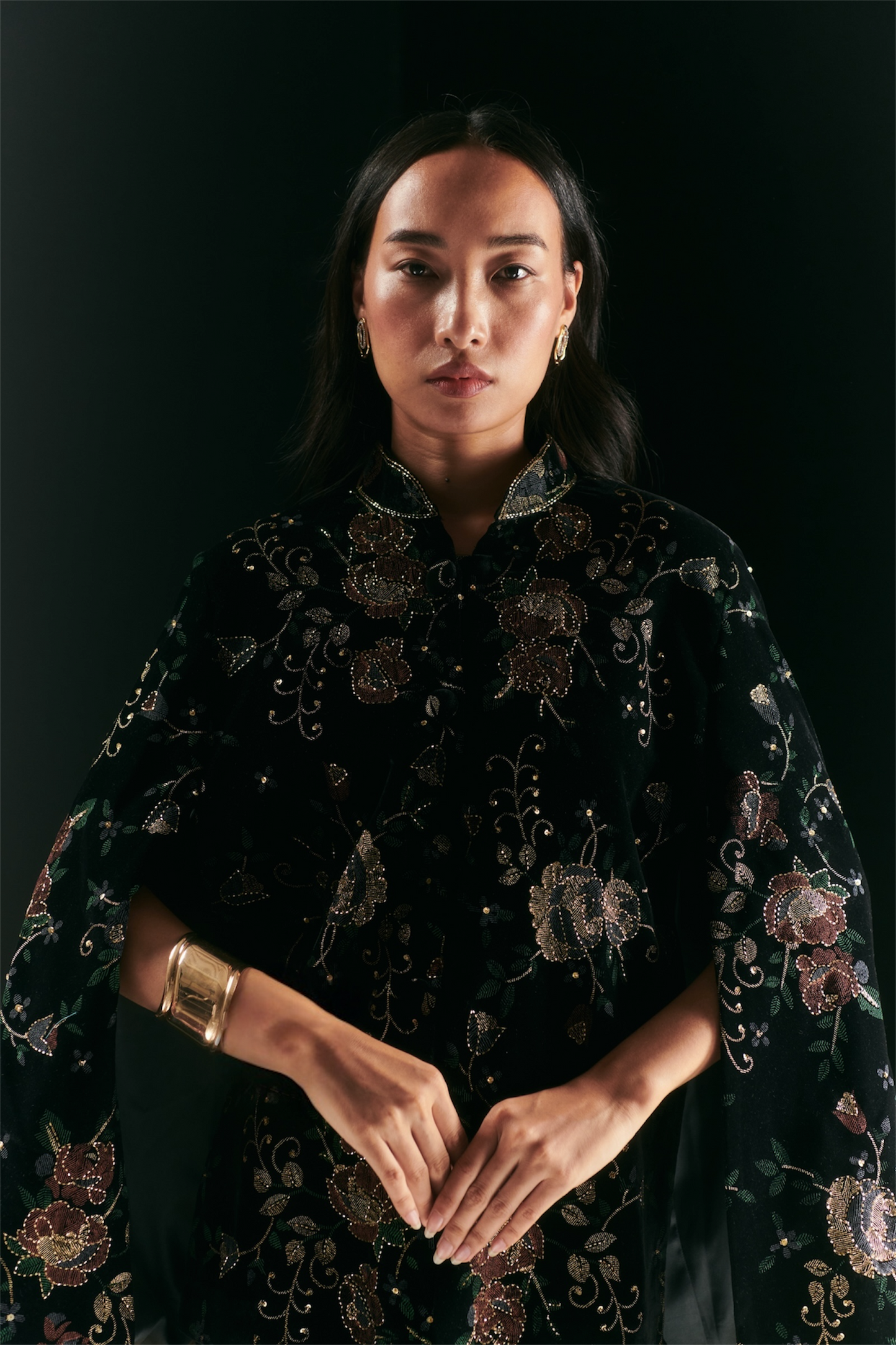 A woman stands against a dark background, wearing a black floral-patterned garment with metallic embroidery. She has long dark hair, gold earrings, and the handcrafted AEQEA TIFFANY CUFF from the Nature's Symphony Collection.